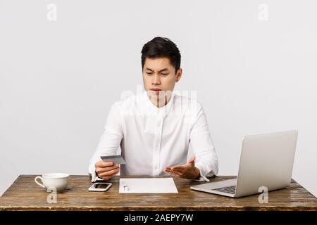 Distressed questioned and frustrated young asian guy sitting office at work, with documents and laptop, looking at credit card and complaining strange Stock Photo