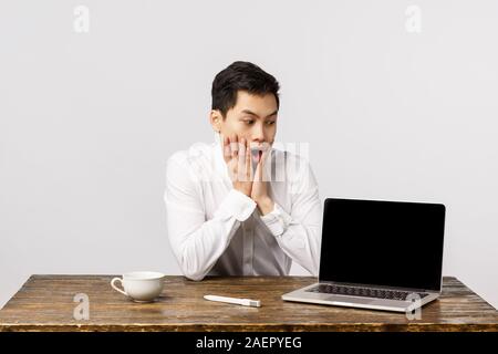 Shocked, speechless, astonished asian office guy sitting desk at work, reacting shook and surprised as looking laptop display, showing something Stock Photo