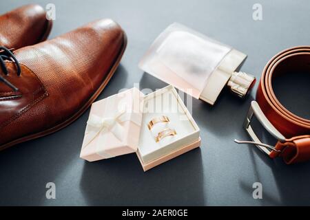 Groom's wedding day preparation accessories. Brown leather shoes, belt, perfume, golden rings in box on grey background. Male fashion Stock Photo