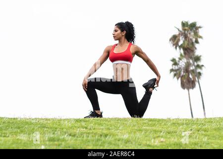 Latina athlete performs quad stretch outdoors wearing fitness leggings and red sports bra with palm trees in background Stock Photo