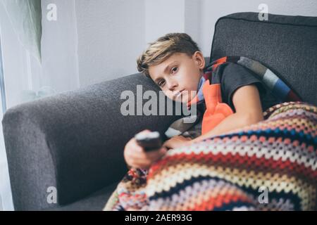 Sick boy watching tv with woolen blanket and hot water bottle. Sad teen with the flu rests at home in a cold winter day. Child with seasonal infection Stock Photo