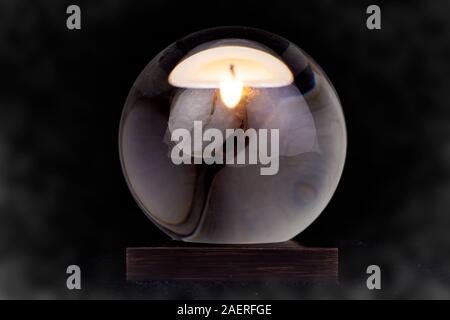 A crystal ball with a candle reflected in it against a black smoky background. Stock Photo