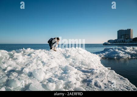 Young smiling woman sitting on the top of sea ice blocks on the coast holding broken piece of ice, with a blue sea and sky and the background Stock Photo