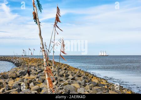 Breakwater or pier and poles with colored rope and plastic waving in the wind and a ship with white sails in the Wadden Sea. Stock Photo