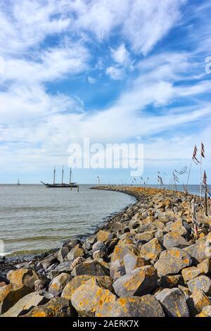 Vertical shot of breakwater or pier and poles with colored rope and plastic waving in the wind and a ship in the Wadden Sea. Stock Photo