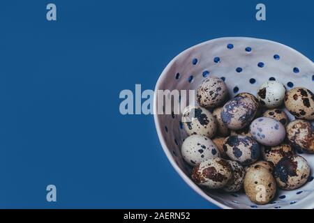 Quail unpainted eggs folded in white ceramic handmade bowl on a blue background Stock Photo