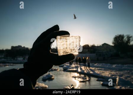 Woman's hand in a black glove holding a broken piece of sea ice against the sun. Person looking through the ice at the sun Stock Photo