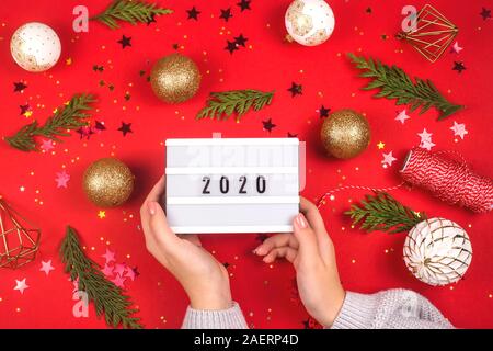 2020 is written on a decorative lamp in female hands among Christmas decorations on a red background. Top view layout. Stock Photo