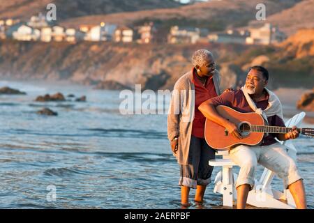 Happy senior woman listening to her husband play an acoustic guitar on a beach. Stock Photo