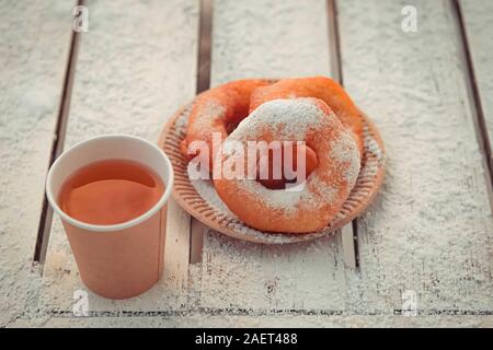 Russian fast food bagel pyshki and paper cup with tea or coffe on wooden table, covered with snow, winter snack in the park. Stock Photo