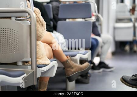 A young lady wearing a sheepskin coat, clear tights, and walking boots sits crossed-legged on a commuter train Stock Photo