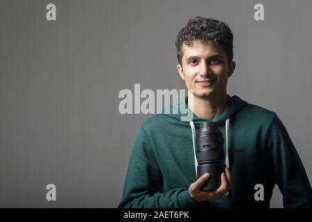Young man wearing green pullover and holding a macro lens on front of his chest in front of dark gray background Stock Photo