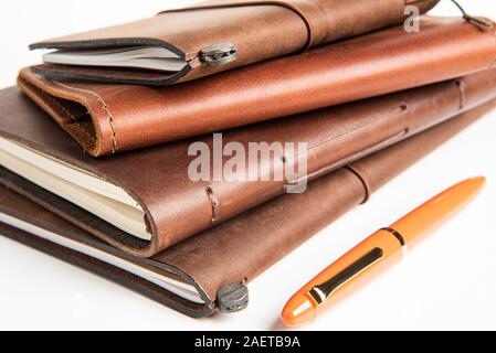 A stack of leather-bound travel writing journals with an orange fountain pen set on plain white background, Stock Photo