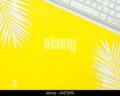 A keyboard on white paper isolated on yellow workplace with white fern leaves on it. Creative workspace. Stock Photo