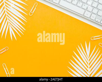 A keyboard on white paper isolated on yellow orange workplace with white fern leaves on it. Creative workspace. Stock Photo