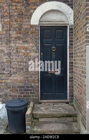 A plastic trash bin is standing by an old door in London, England. The brick wall has seen better days. Stock Photo