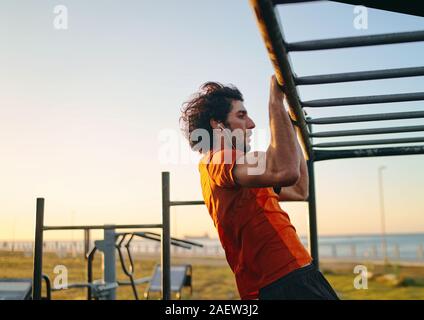 Side view of a muscular fit young man with earphones in his ears doing pull-ups at the outdoor gym in the park Stock Photo