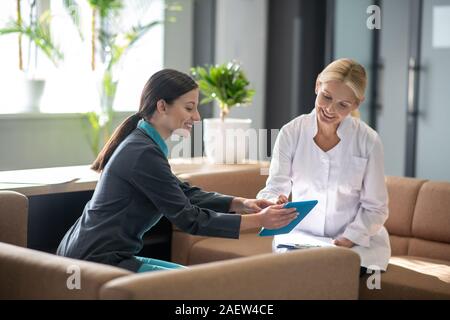 Dark-haired young female intern showing something on tablet to her colleague Stock Photo