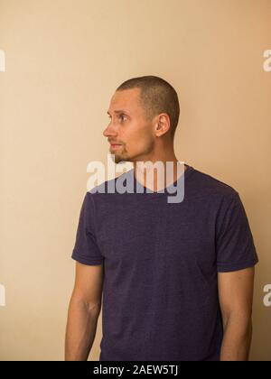Attractive brutal bearded guy poses in blank purple t-shirt on light biege background. Stock Photo