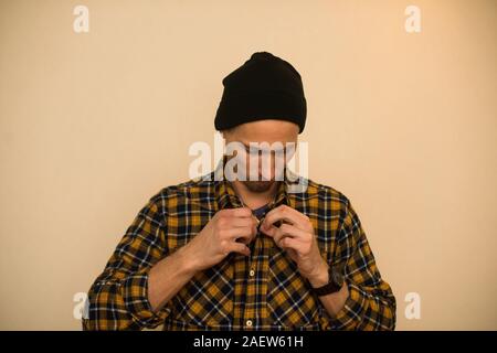 Young man, model of fashion, wearing a plaid shirt with a biege blind behind him. Guy in black hat with beard in casual style Stock Photo