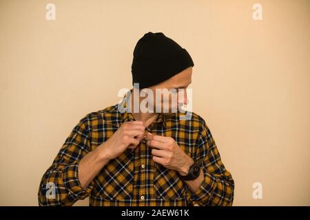 Young man, model of fashion, wearing a plaid shirt with a biege blind behind him. Guy in black hat with beard in casual style Stock Photo