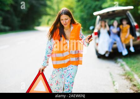 Woman in orange reflective vest installing red warning triangle on the road, children and broken car in background Stock Photo