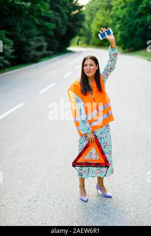Woman with red warning triangle in orange reflective vest is catching car on the forrest road Stock Photo