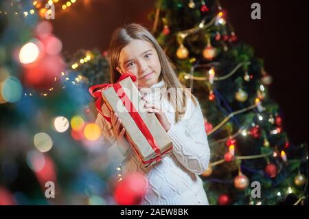 Beautiful girl in the room near the festive Christmas tree holds a gift wrapped in red ribbon. Holidays. Christmas and New Year. Stock Photo