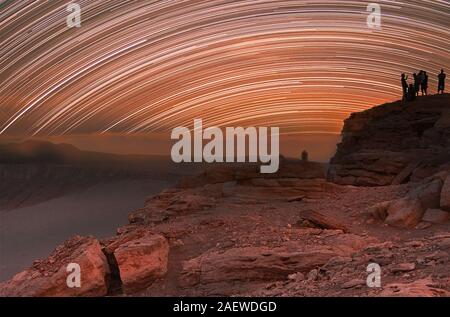 Young people is on the rock. Night glowing stars trails in the sky.  Natural stony desert background. Long exposure Stock Photo