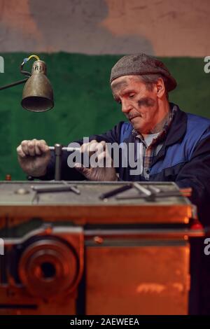 Serious senior man in work wear turning the lever while working at the lathe in the factory Stock Photo