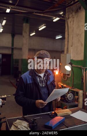 Senior manual worker in work wear sitting at his workplace and reading the document in the factory Stock Photo