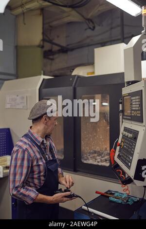 Mature manual worker in work wear watching the work of machine and operating it with control panel Stock Photo