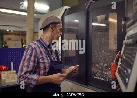 Mature engineer holding digital tablet and watching for the process of work while standing near the lathe in the factory Stock Photo
