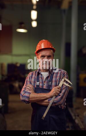 Portrait of senior manual worker in work helmet holding measuring equipment and looking at camera while standing in the factory Stock Photo