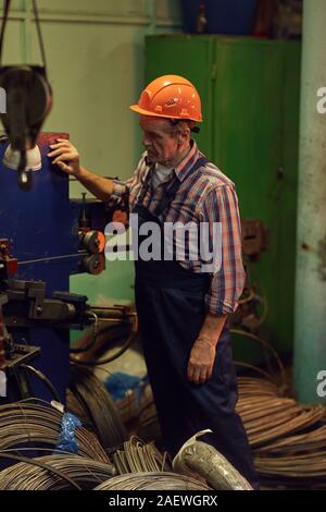 Mature engineer in work helmet standing at the lathe among cables and watching for the work of lathe Stock Photo