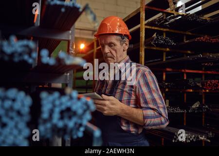 Senior foreman in work helmet standing near the shelves with metal pipes and using digital tablet at warehouse Stock Photo