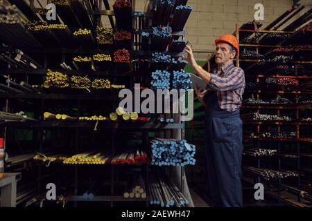 Manual worker in work helmet using digital tablet and checking the metal pipes on the shelves in the factory Stock Photo