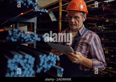 Serious mature foreman in work helmet working online on tablet pc while standing near the shelves with metal pipes Stock Photo