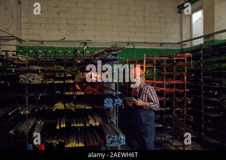 Serious senior man in work helmet checking the online order on tablet pc while standing near the metal pipes in warehouse Stock Photo