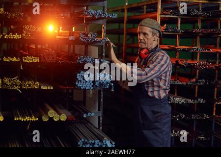 Manual worker in overalls looking at metal pipes on the shelves and using digital tablet in warehouse Stock Photo