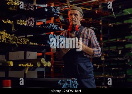 Mature manual worker in overalls using digital tablet and looking at colorful metal pipes on shelves Stock Photo