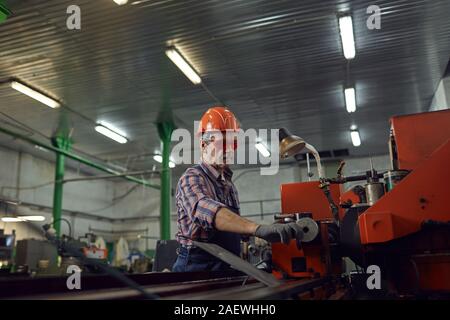 Mature manual worker wearing protective eyeglasses standing at lathe holding the lever and controlling the machine Stock Photo