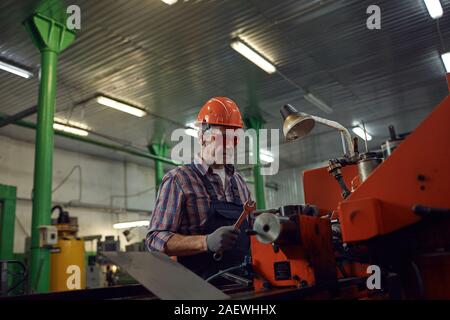 Mature mechanic in protective eyeglasses and in work helmet repairing the lathe with work tool Stock Photo