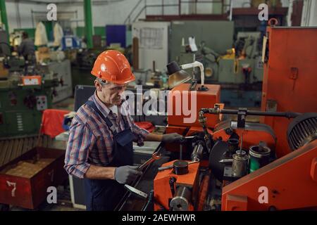 Mature worker in work helmet using work tools while repairing the lathe in the plant Stock Photo
