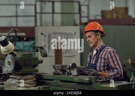 Serious mature manual worker in work helmet sitting at the lathe and using digital tablet Stock Photo