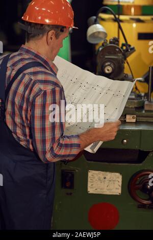 Rear view of mature foreman in work helmet standing and examining the blueprint before work in the plant Stock Photo