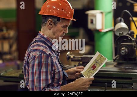 Mature manual worker in work helmet looking at plan in his hands before his work on lathe Stock Photo