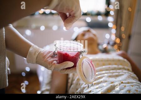 Cropped therapist pouring pink sea salt scrub in jar. SPA therapist hands with latex gloves pouring scrub into glass jar with pink salt next to lying Stock Photo