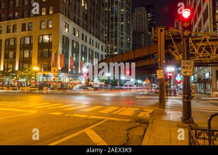 Chicago, IL - Circa 2019: Night time long exposure of busy downtown Chicago intersection roadway under loop train track. Red traffic light on corner d Stock Photo