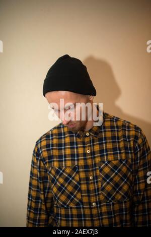 Young man, model of fashion, wearing a plaid shirt with a biege blind behind him. Guy in black hat with beard in casual style. Stock Photo